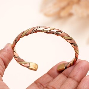 May include: A cuff bracelet constructed from three twisted metal strands in copper, brass, and silver tones. The open-ended design features smooth, rectangular metal end caps at each terminal, showcasing a multi-toned metallic finish throughout.