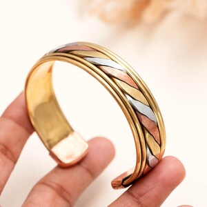 May include: A cuff bracelet featuring a tri-color design with intertwined strands of gold, silver, and copper-toned metal. The band has a polished finish with smooth edges and a wide, open-ended structure that allows for easy adjustment on the wrist.