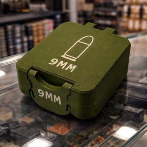 Puede incluir: Una funda verde oliva con un gráfico blanco de una bala y el texto "9MM" en blanco. La funda rectangular tiene una tapa con bisagras y un cierre. La funda está sobre una superficie de vidrio.