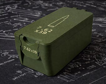 May include: An olive green rectangular box with a hinged lid. The box features the text "7.62 X39" and a bullet graphic in white. The box is on a black surface with a white schematic design.