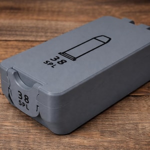 May include: A gray ammunition box with the text "38 SPL" and a graphic of a bullet. The box is rectangular with a hinged lid and a latch. The box is sitting on a wooden surface.