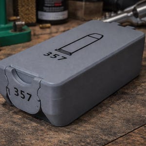 May include: A grey plastic ammunition storage box with the number "357" printed on the front and top. The top of the box has a graphic of a bullet. The box is on a wooden surface.