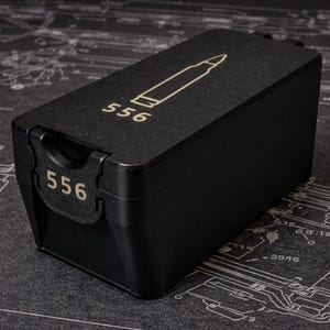 Puede incluir: Un contenedor rectangular negro con el número "556" y un gráfico de bala en dorado. El contenedor está sobre una superficie oscura con un patrón de dibujo técnico.