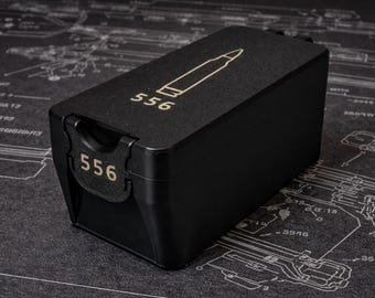 May include: A black rectangular container with the number "556" and a bullet graphic in gold. The container is on a dark surface with a technical drawing pattern.