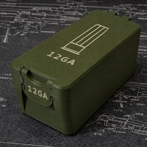 May include: An olive green rectangular container with the text "12GA" printed on the front and top. The top of the container features a white graphic of shotgun shells. The container is on a dark surface.