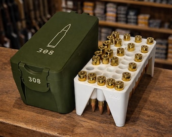 May include: An olive green ammunition box and a white ammunition holder with gold-colored bullets. The box has the number "308" and a bullet graphic. The holder is filled with bullets and is on a wooden surface.