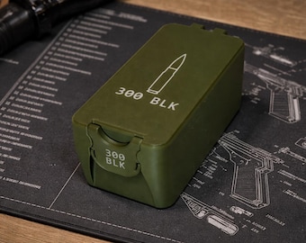 May include: An olive green ammunition box with the text "300 BLK" and a bullet graphic. The box is rectangular with a hinged lid and a latch. The box is on a black surface with a gun schematic.