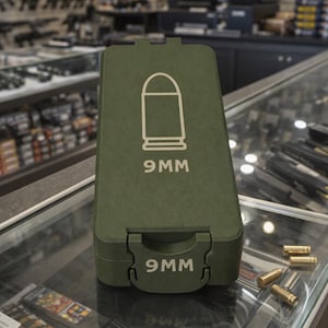 May include: An olive green ammunition case with a white outline of a bullet and the text "9MM" printed on the front. The case is on a glass surface with several loose bullets.