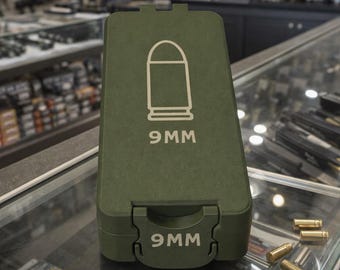 May include: An olive green ammunition case with a white outline of a bullet and the text "9MM" printed on the front. The case is on a glass surface with several loose bullets.