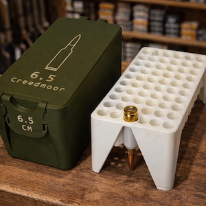Könnte beinhalten: Eine olivgrüne Munitionskiste und ein weißer Patronenhalter liegen auf einer Holzoberfläche. Auf der Kiste steht "6.5 Creedmoor" und "6.5 CM". Der Patronenhalter hat eine einzelne Patrone.