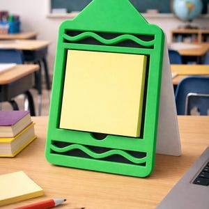 May include: A green crayon-shaped desk organizer holding a stack of yellow sticky notes. The organizer sits on a wooden desk with pencils, more sticky notes, and a laptop. A chalkboard with math equations is in the background.