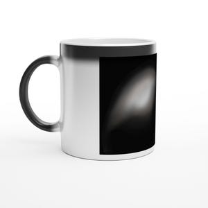 Könnte beinhalten: Weiße Keramiktasse mit schwarzem Henkel und einem schwarz-weißen Design an der Seite. Das Design scheint ein Farbverlauf von Schwarz und Weiß zu sein, der eine glatte, abstrakte Form erzeugt. Die Tasse steht auf einer weißen Oberfläche.