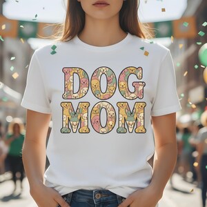 May include: A white t-shirt featuring the text "DOG MOM" in large, colourful letters. The letters are filled with illustrations of different dogs in shades of yellow, pink, and green. The shirt has short sleeves and is likely made of cotton.