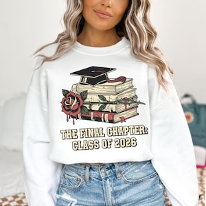 May include: White sweatshirt featuring a graphic of a graduation cap atop a stack of books, a red rose, and the text "THE FINAL CHAPTER: CLASS OF 2026." The design is in a vintage style. The sweatshirt is a crew neck.