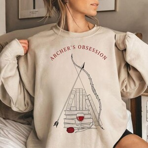 May include: Beige sweatshirt with the text "ARCHER'S OBSESSION" in red. The graphic features a bow and arrow, books, a wine glass, and a red rose. The design is in black and red.