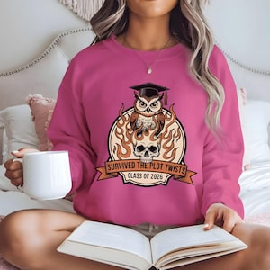 May include: A magenta sweatshirt featuring a graphic of an owl wearing a graduation cap, flames, and a skull. The text reads "Survived the Plot Twists Class of 2026". The sweatshirt is a crew neck style.