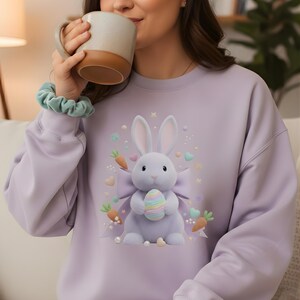 Puede incluir: Una sudadera lila con una ilustración 3D de un conejito sosteniendo un huevo de Pascua colorido. El diseño incluye zanahorias, corazones y perlas. La sudadera es de estilo casual y cómodo.