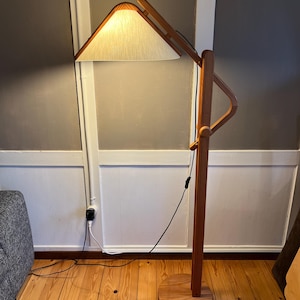 Puede incluir: Una lámpara de pie de madera con una pantalla triangular. La lámpara tiene una pantalla de tela de color claro y una estructura de madera. La base es redonda y la lámpara es de madera clara. La lámpara está enchufada a una toma de corriente.