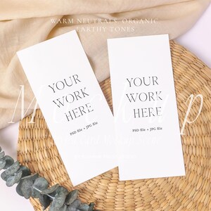 Op de afbeelding: Twee verticale witte rechthoekige kaarten met de zwarte tekst YOUR WORK HERE en PSD file + JPG file. De kaarten liggen op een ronde gevlochten strooien mat naast een beige stoffen doek en een groene eucalyptus tak met kleine blaadjes.