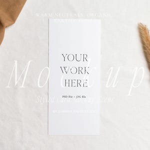 May include: A styled card mockup scene featuring a white card with the text "YOUR WORK HERE". The card is placed on a white surface with a brown fabric and a dried plant stem. The scene has a neutral, organic aesthetic.