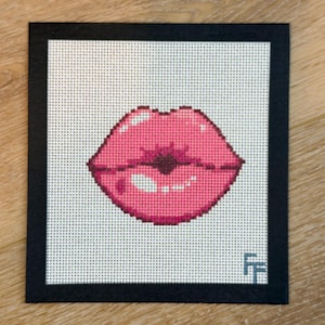 May include: A framed cross-stitch artwork featuring a pair of pink lips. The lips are rendered in a pixelated style, with varying shades of pink and a glossy effect. The artwork is set against a white background and framed in black.