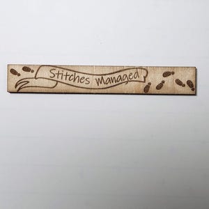 May include: A rectangular wooden sign with the text "Stitches Managed" in a banner design. The sign has footprints on each end. The wood has a light brown colour.