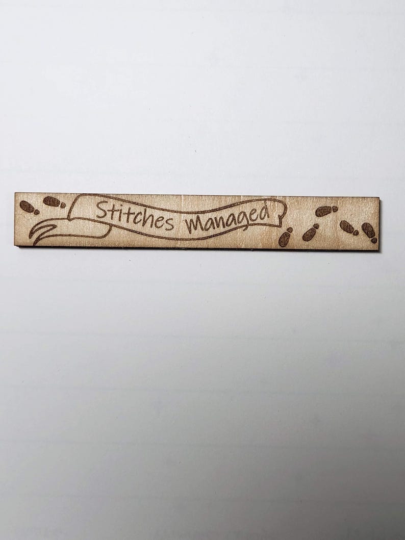 May include: A rectangular wooden sign with the text "Stitches Managed" in a banner design. The sign has footprints on each end. The wood has a light brown colour.