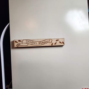 May include: A wooden magnet displaying the words "Stitches Managed" within a banner, decorated with tiny footprints. The magnet is attached to a white surface.