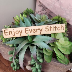 May include: A wooden planter box filled with various green succulents. A wooden sign with the words "Enjoy Every Stitch" is placed in the center of the arrangement. The succulents have different shapes and shades of green.
