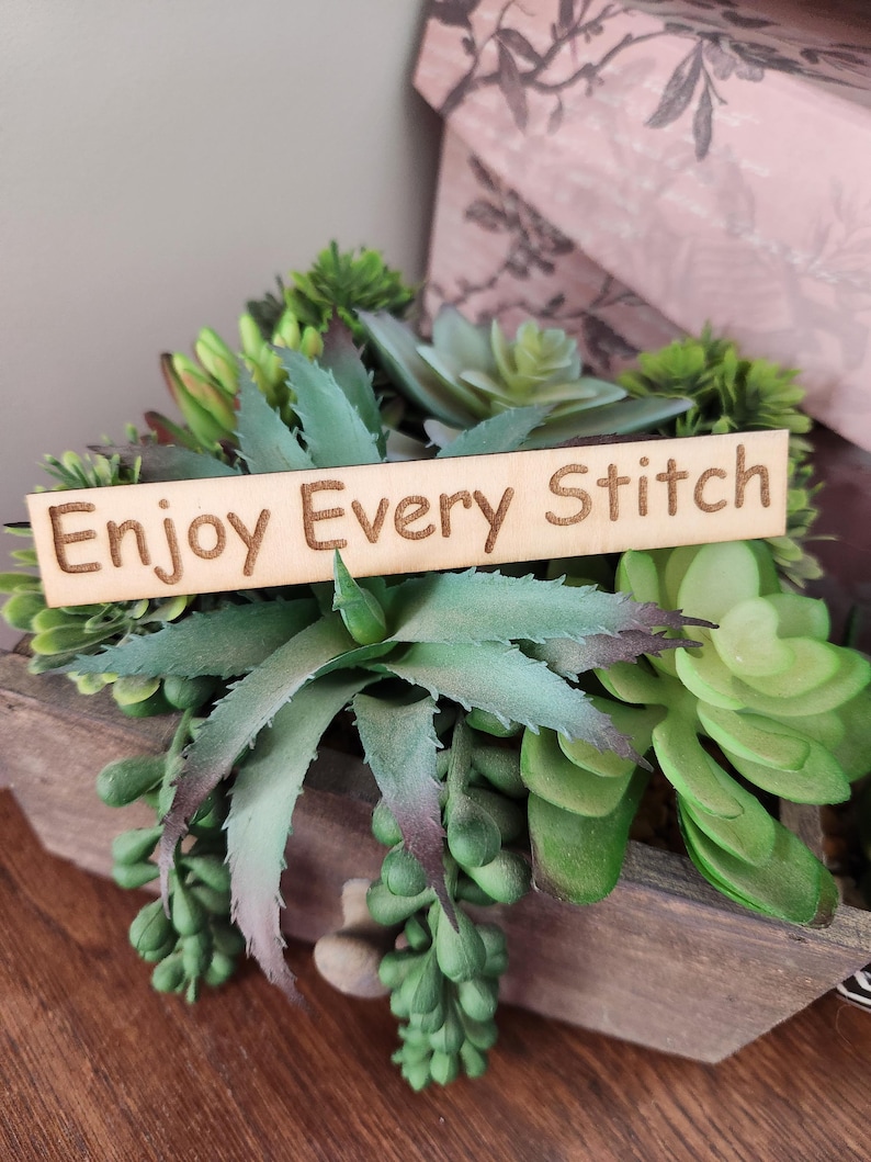 May include: A wooden planter box filled with various green succulents. A wooden sign with the words "Enjoy Every Stitch" is placed in the center of the arrangement. The succulents have different shapes and shades of green.