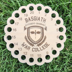 May include: A round, wooden thread organizer with 20 numbered holes around the edge. The center features the text "BASGIATH Stitcher's Quadrant War College" and a crest design. The organizer is on a bed of green grass.