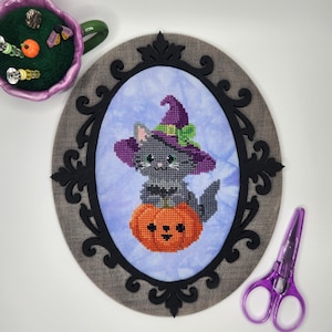 May include: A cross-stitch embroidery of a gray kitten wearing a purple witch hat, sitting on an orange jack-o'-lantern. The piece is set in an oval black frame with a gray background, accompanied by a purple pincushion and purple scissors.