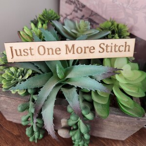 May include: A wooden planter box filled with assorted green succulents. A wooden sign with the text "Just One More Stitch" is placed in front of the plants. The box is on a wooden surface.