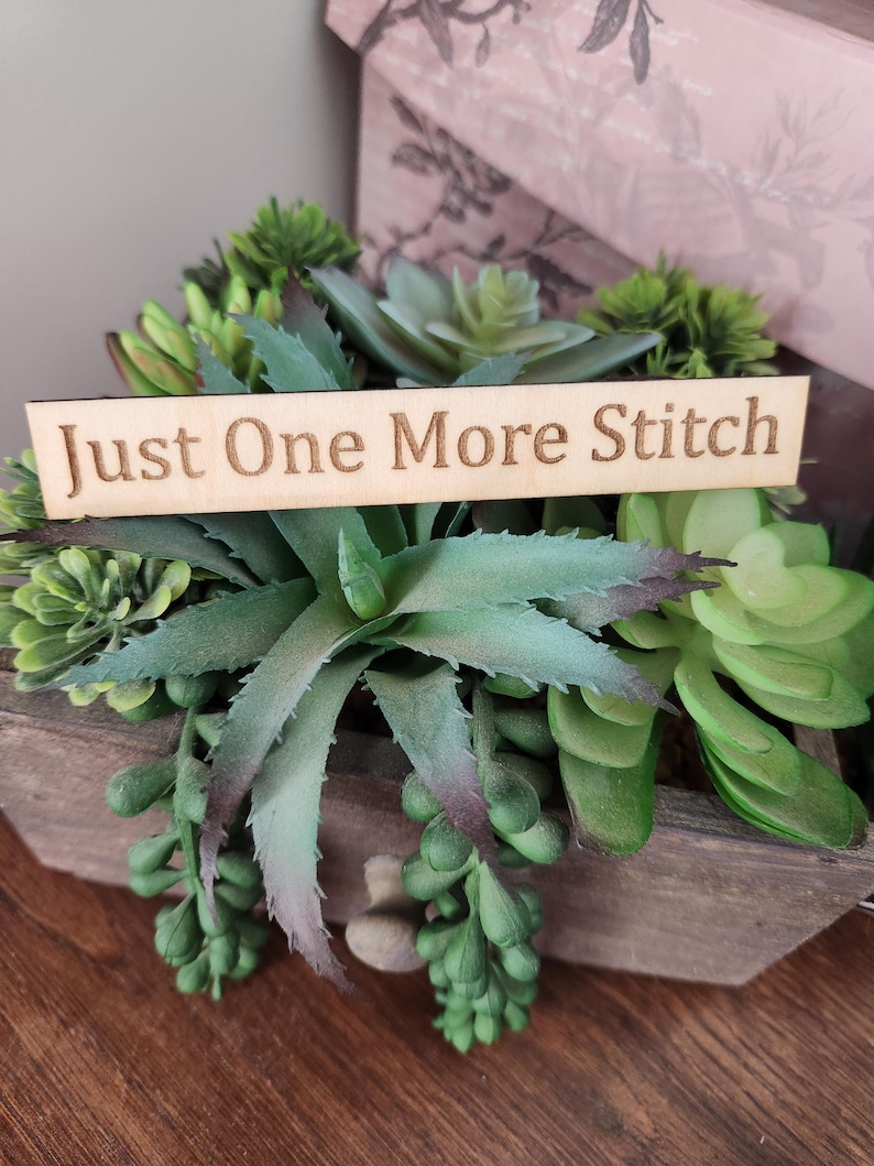 May include: A wooden planter box filled with assorted green succulents. A wooden sign with the text "Just One More Stitch" is placed in front of the plants. The box is on a wooden surface.