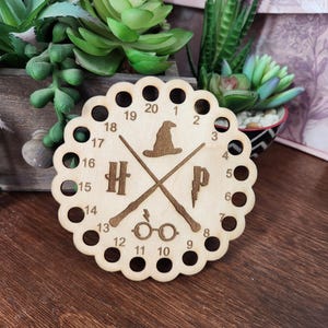 May include: A wooden Harry Potter-themed clock with a scalloped edge and twenty numbered holes. The clock features crossed wands, a sorting hat, glasses, and the letters "H" and "P". The clock is made of light-colored wood.