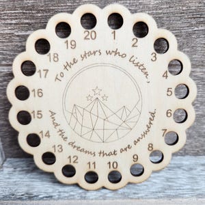 May include: A round, wooden craft with a scalloped edge and twenty numbered holes. The center features a mountain design and the text "To the stars who listen, And the dreams that are answered."