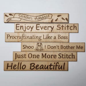 May include: Wooden laser-cut signs with various phrases, including "Stitches Managed," "Enjoy Every Stitch," and "Hello Beautiful." The signs are light brown with dark brown engraved text and designs, perfect for crafters.