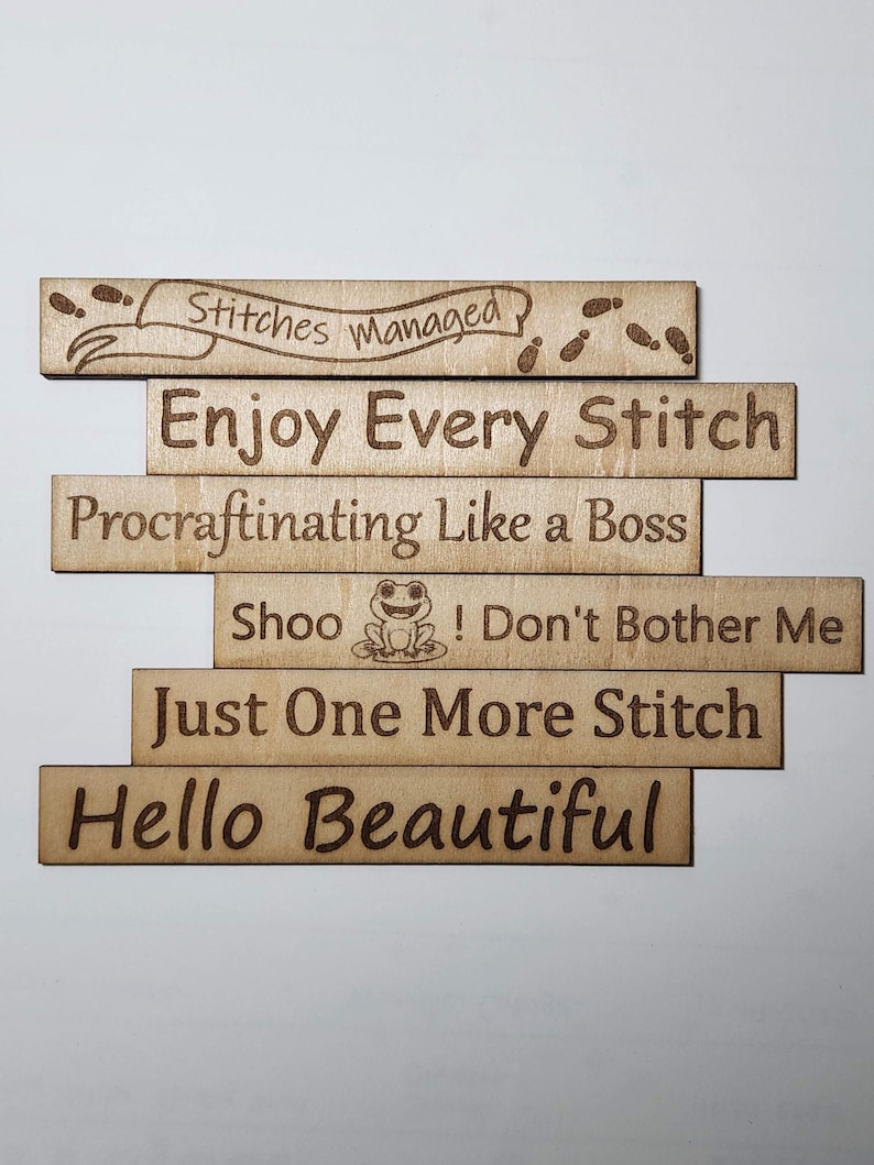 May include: A collection of laser-cut wooden signs, each bearing a different phrase such as "Enjoy Every Stitch" and "Procraftinating Like a Boss." The light brown wood has dark brown engraved text, ideal for craft enthusiasts.