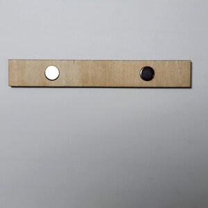 May include: A light brown wooden bar with two circular magnets attached. One magnet is silver, and the other is dark grey. The bar is rectangular and appears to be made of unfinished wood.