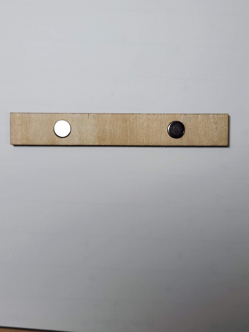 May include: A light brown wooden bar with two circular magnets attached. One magnet is silver, and the other is dark grey. The bar is rectangular and appears to be made of unfinished wood.