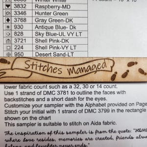 May include: A light brown wooden craft piece with the words "Stitches Managed" in a banner design, and small footprint illustrations. It sits on white paper with colour codes and instructions for a cross-stitch project.