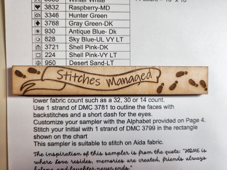 May include: A light brown wooden craft piece with the words "Stitches Managed" in a banner design, and small footprint illustrations. It sits on white paper with colour codes and instructions for a cross-stitch project.