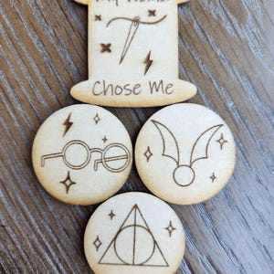 May include: Wooden Harry Potter-themed embellishments. The top piece reads "My wand chose me" and is shaped like a spool. Below are three round pieces with etched designs: glasses, a snitch, and the Deathly Hallows symbol. Sparkles and lightning bolts are also etched.