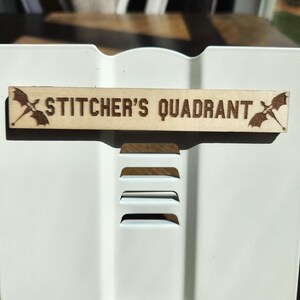 May include: A rectangular light-colored wooden sign with the text STITCHER'S QUADRANT laser-engraved in the center. The sign features a small winged creature illustration on both the left and right sides of the text, positioned horizontally.