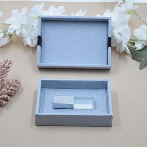 Puede incluir: Dos bandejas de tela gris, una vacía y otra con un pequeño espejo rectangular y un bloque rectangular transparente. Las bandejas son rectangulares con un borde elevado y un interior de tela azul claro. El fondo incluye flores blancas y una superficie beige.