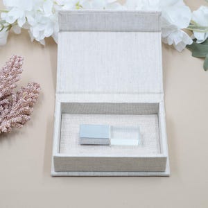 May include: An open, rectangular, linen-covered box with a light blue USB drive inside. The box is beige and has a hinged lid. White flowers and pink coral-like branches are in the background.