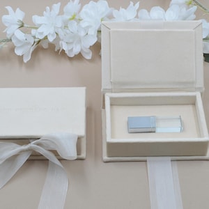 May include: A cream-colored USB drive rests inside an open, matching box. The box is lined with soft fabric and has a clear lid. A second closed box with a ribbon is visible. White flowers and a ribbon are in the background. The text "OUR WEDDING DAY" is embossed on the closed box.