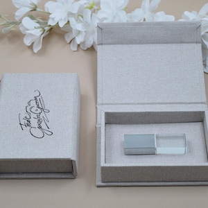 May include: A light gray linen-textured presentation box with a magnetic lid featuring black cursive text that reads Fredy Gonzalez Photography. Inside the open box sits a rectangular silver and clear glass USB flash drive.