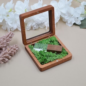May include: A square wooden box with a hinged glass lid featuring the text Retratos. Inside, a rectangular USB flash drive with a wooden cap rests on a bed of bright green preserved moss. The box has a smooth, dark brown wood finish.
