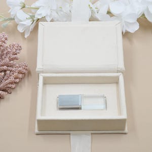 Puede incluir: Caja de tela blanquecina abierta con una unidad USB plateada y transparente en su interior. La caja tiene un cierre de cinta y está forrada con un material suave. Flores blancas y ramitas decorativas rosas en el fondo.